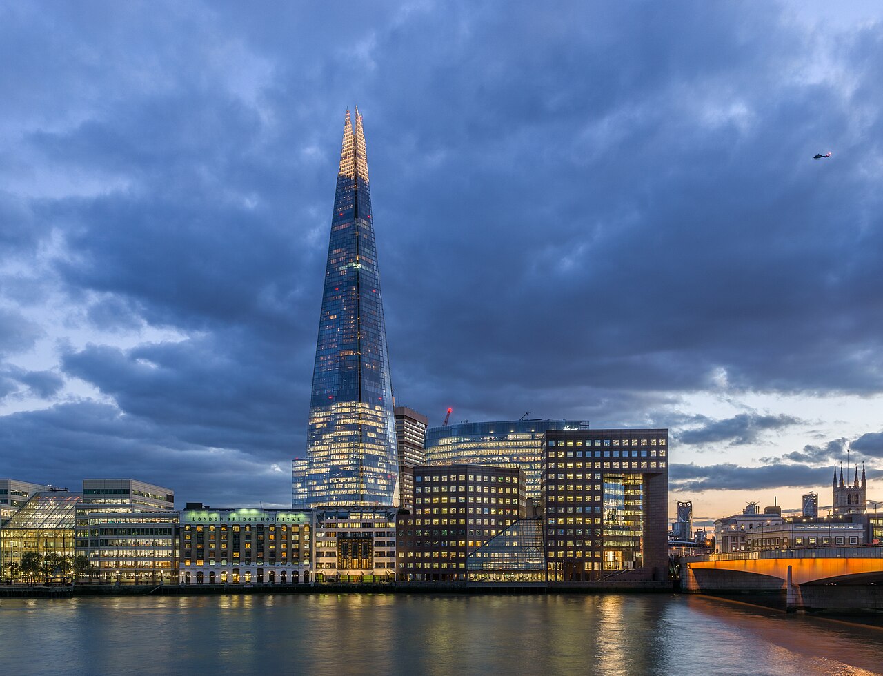 A tall glass tower tapering to a point, lit from inside, rising above lower riverside buildings under a heavy blue evening sky.