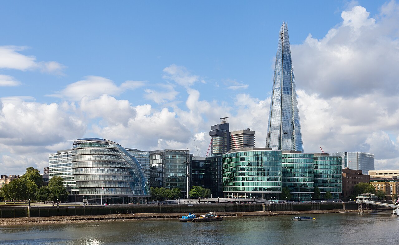 A rounded glass building and glass office blocks on a riverbank, with a pointed glass skyscraper rising behind them under white clouds.