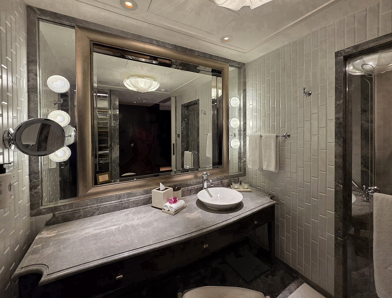 A dark stone vanity with a round basin under a wide framed mirror, a lit magnifying mirror and a towel on a rail beside it.