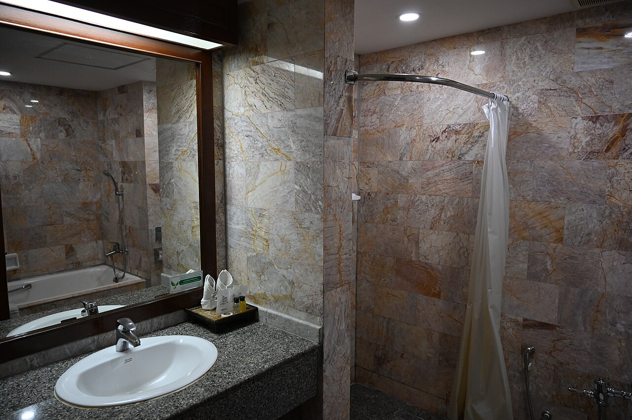 A white basin set in a speckled granite counter below a large mirror, next to a shower with a curved rail and white curtain.