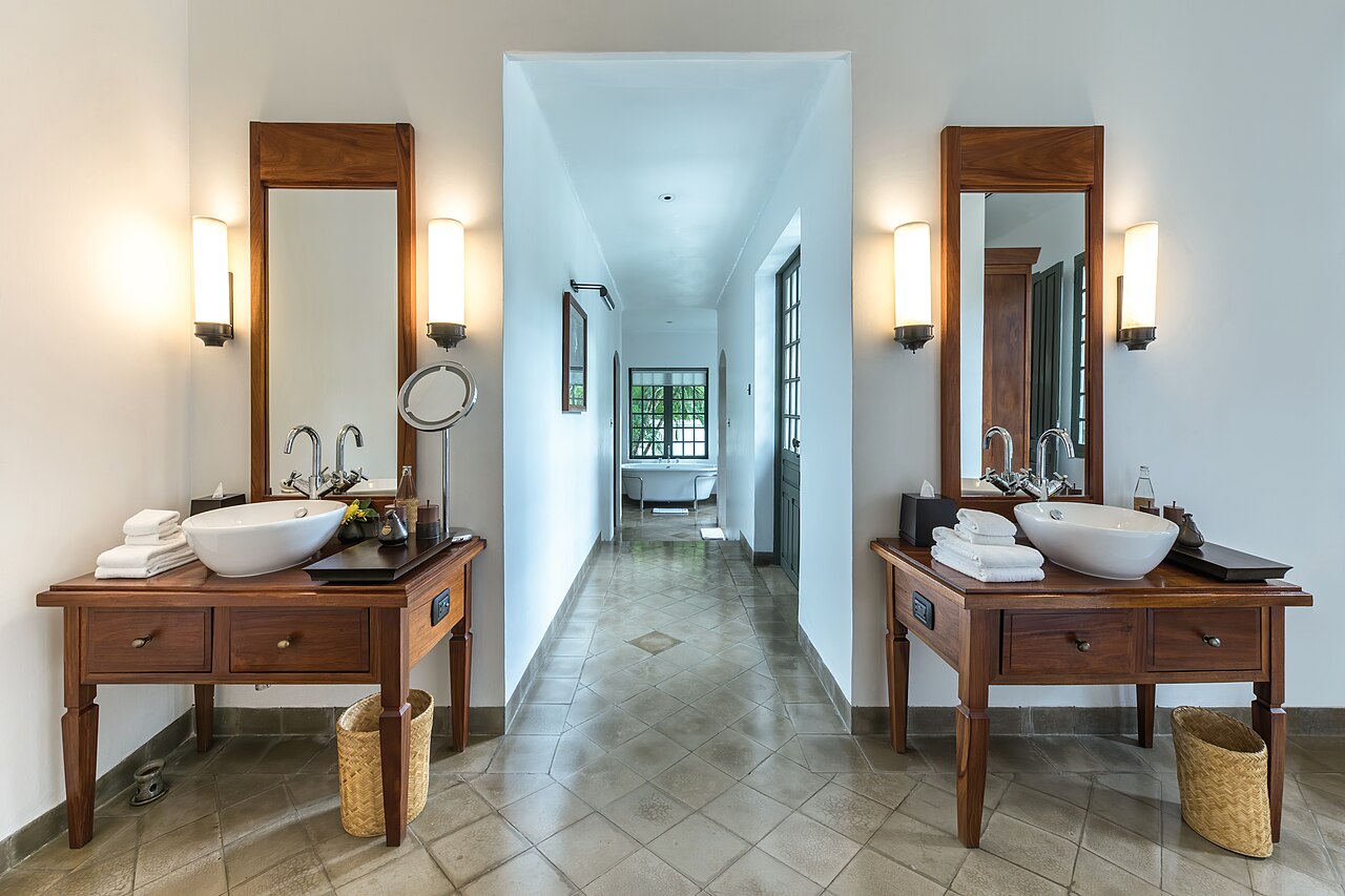 Two matching wooden washstands with bowl basins, tall mirrors and folded white towels face each other across a tiled corridor.