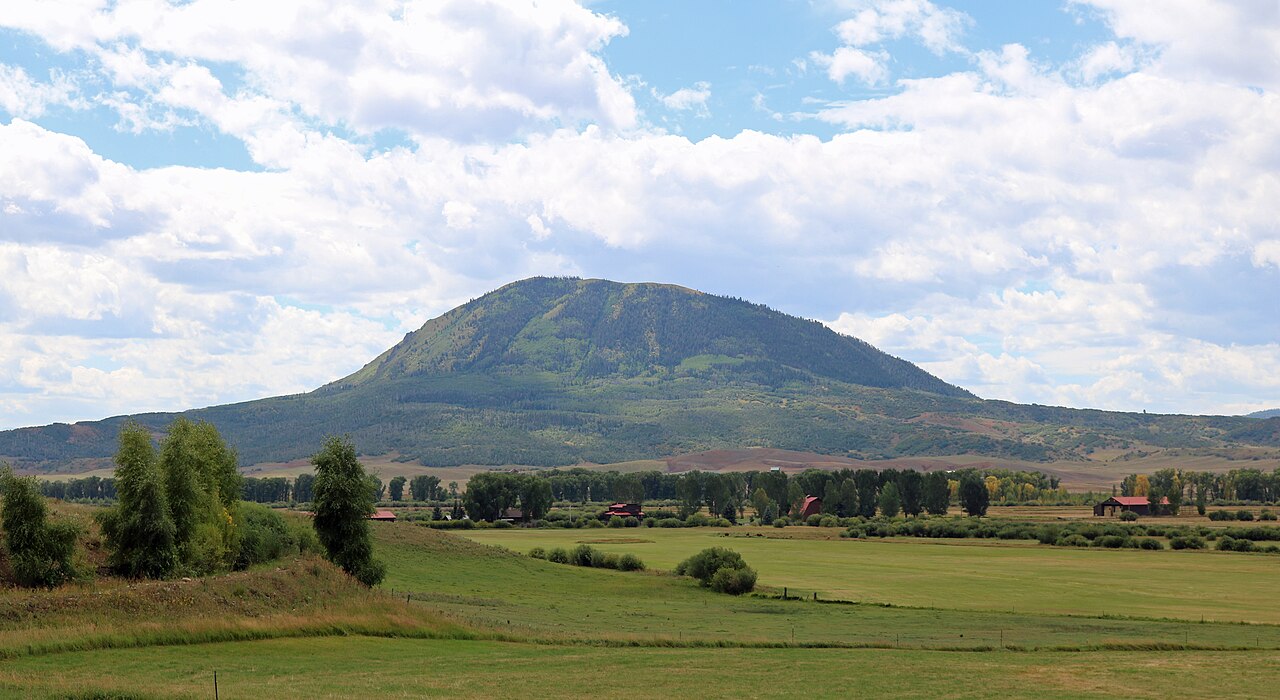 A rounded green mountain rising alone above flat meadows and scattered trees under a sky of white clouds.