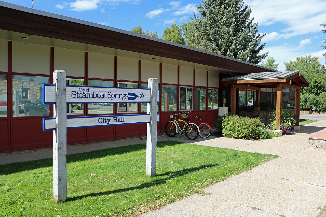A low red and cream building with a timber porch behind a white post sign, with a lawn and bicycles in front.