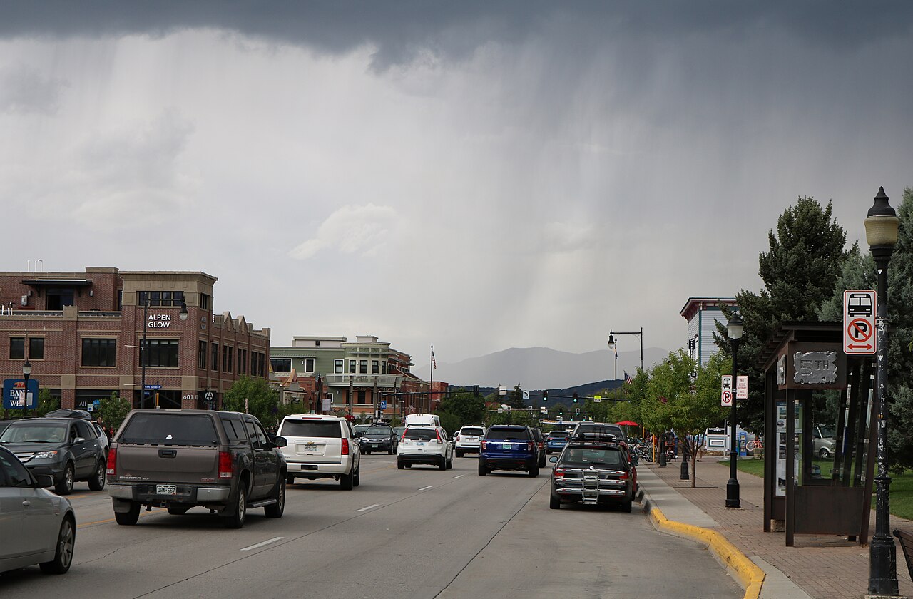 Cars and pickup trucks on a wide street between low brick buildings, with dark rain clouds over the hills ahead.