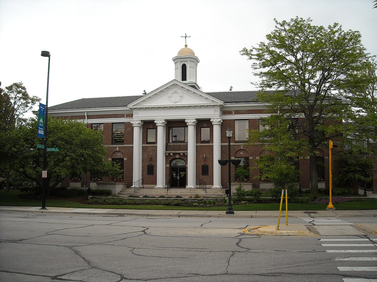 A red brick building with a white columned portico and a small white cupola on its roof, seen across a street beside a large tree.