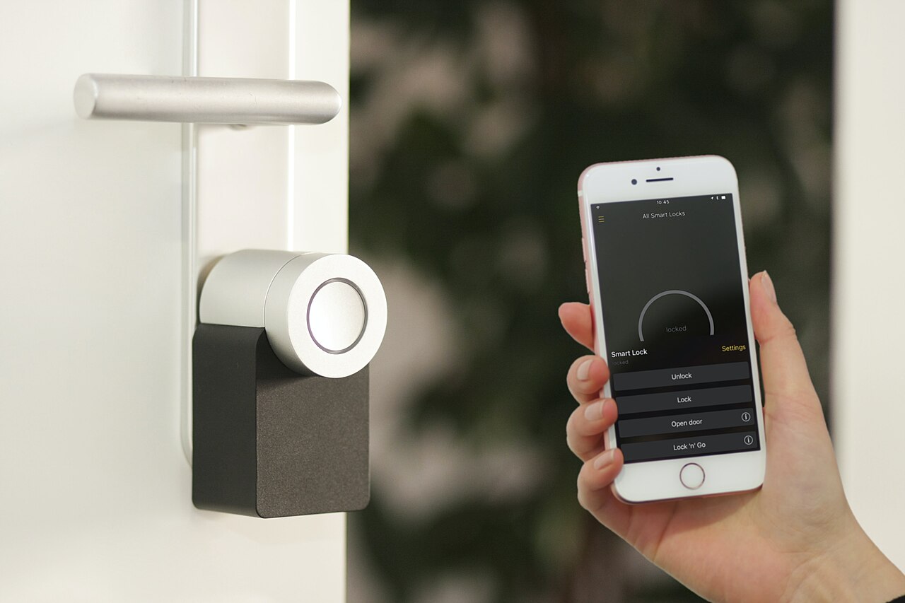 A hand holds a phone showing a lock app next to a white door with a round motorised lock unit below the handle.