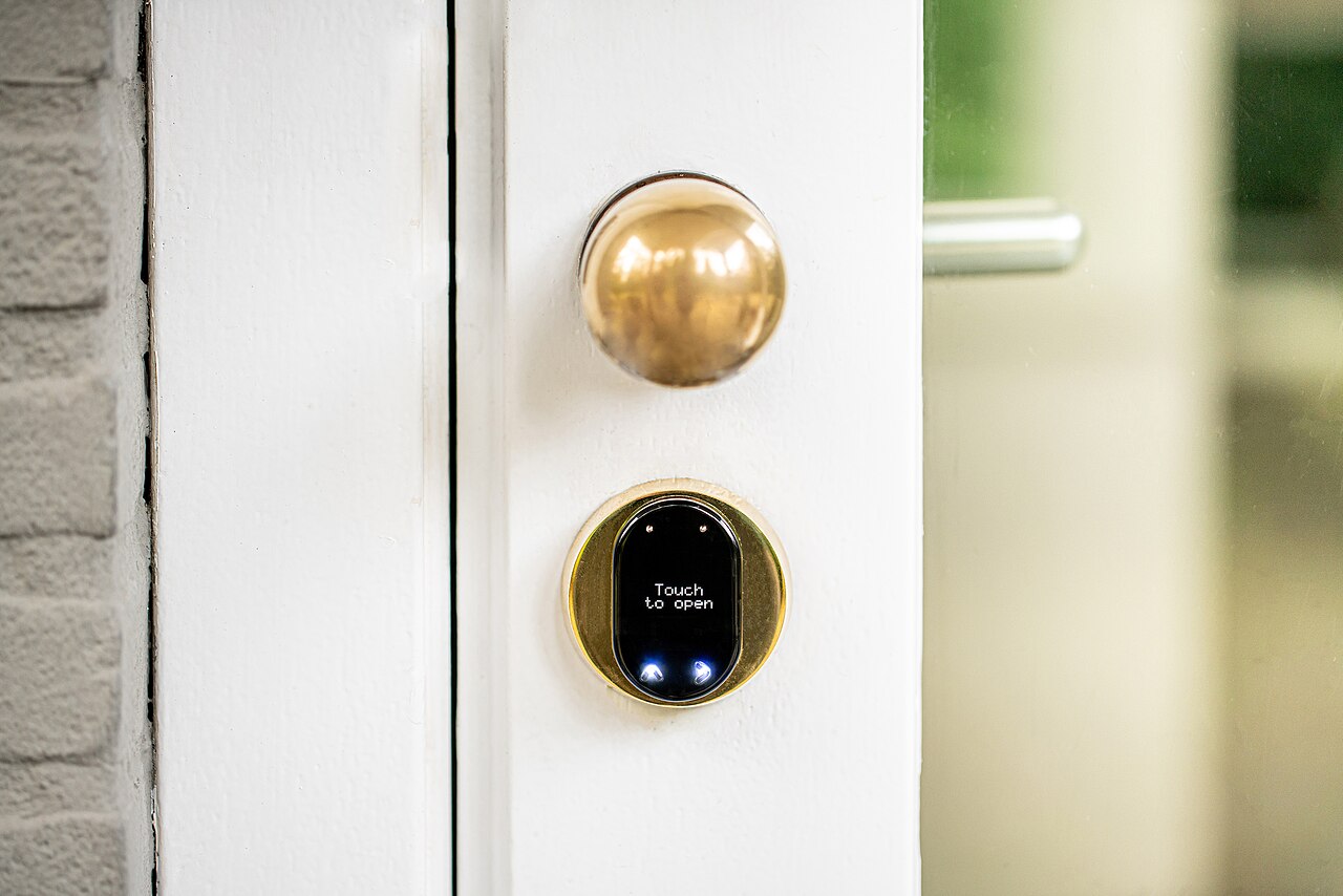 A white door with a brass knob above a round brass lock plate that has a small black touch panel instead of a keyhole.