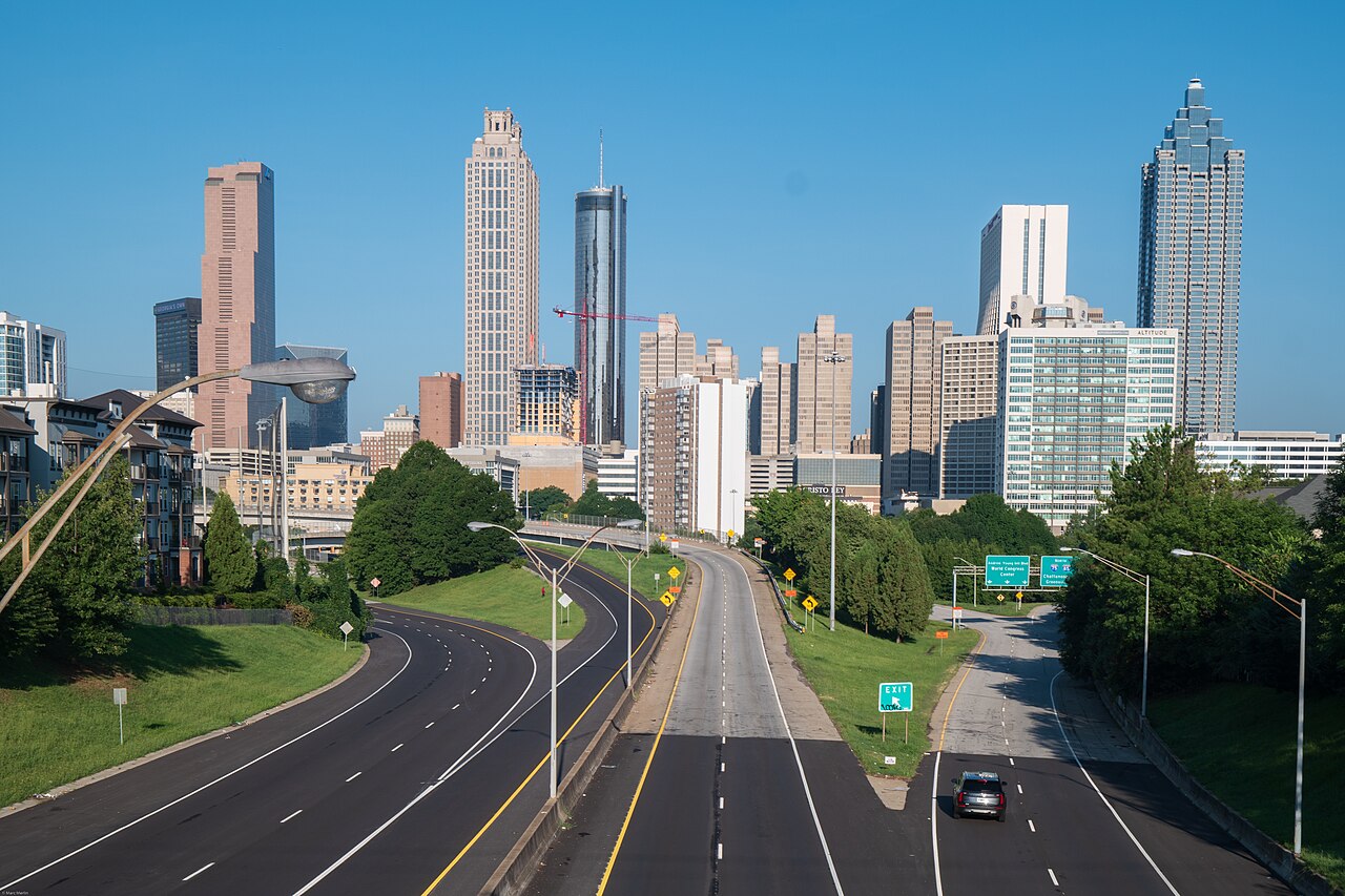 A highway runs between green banks toward a group of downtown towers under a clear blue sky.