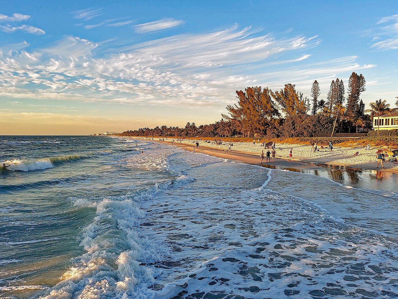 Waves roll onto a long sandy beach lined with trees and low houses in warm evening light.