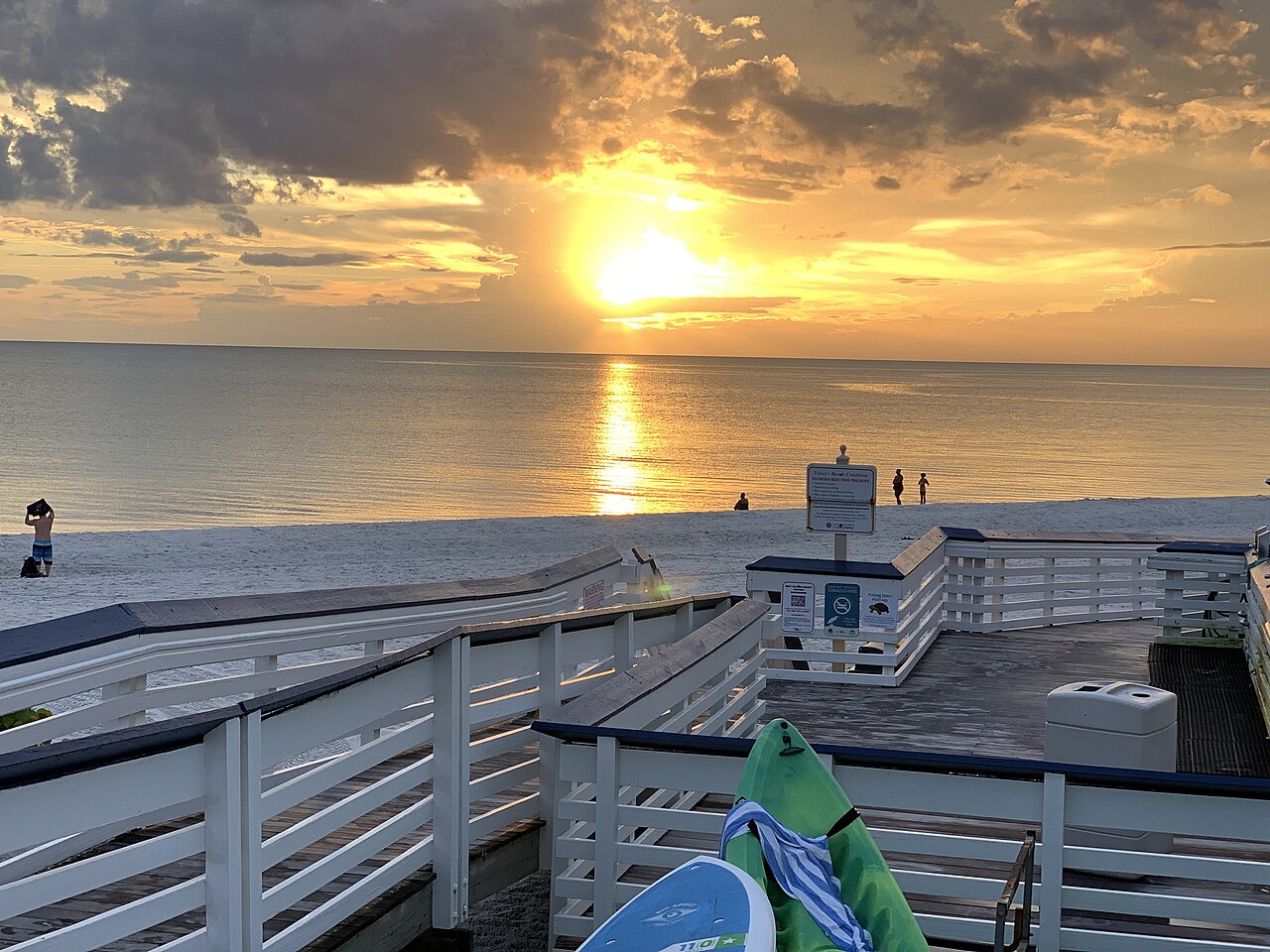 The sun sets through clouds over a calm sea, with a white wooden boardwalk and beach in the foreground.
