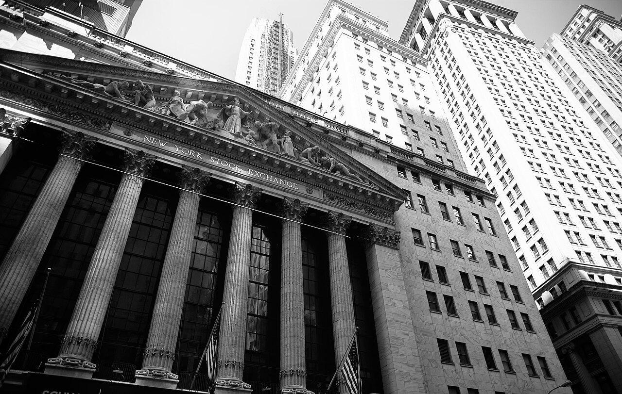 A black and white view looking up at a row of tall stone columns and a carved pediment, with office towers behind.