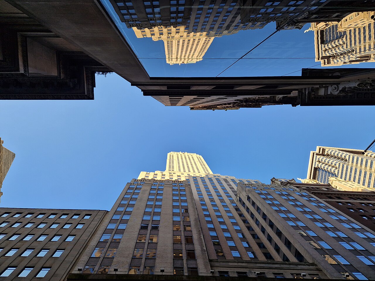 A view straight up between tall stone and glass towers to a patch of blue sky, the upper floors lit gold by the sun.