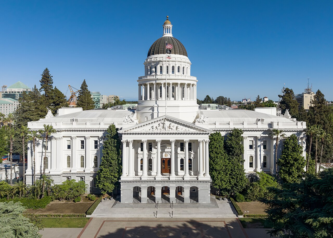 A white domed capitol building with a columned portico, seen from above its front lawn under a clear blue sky.