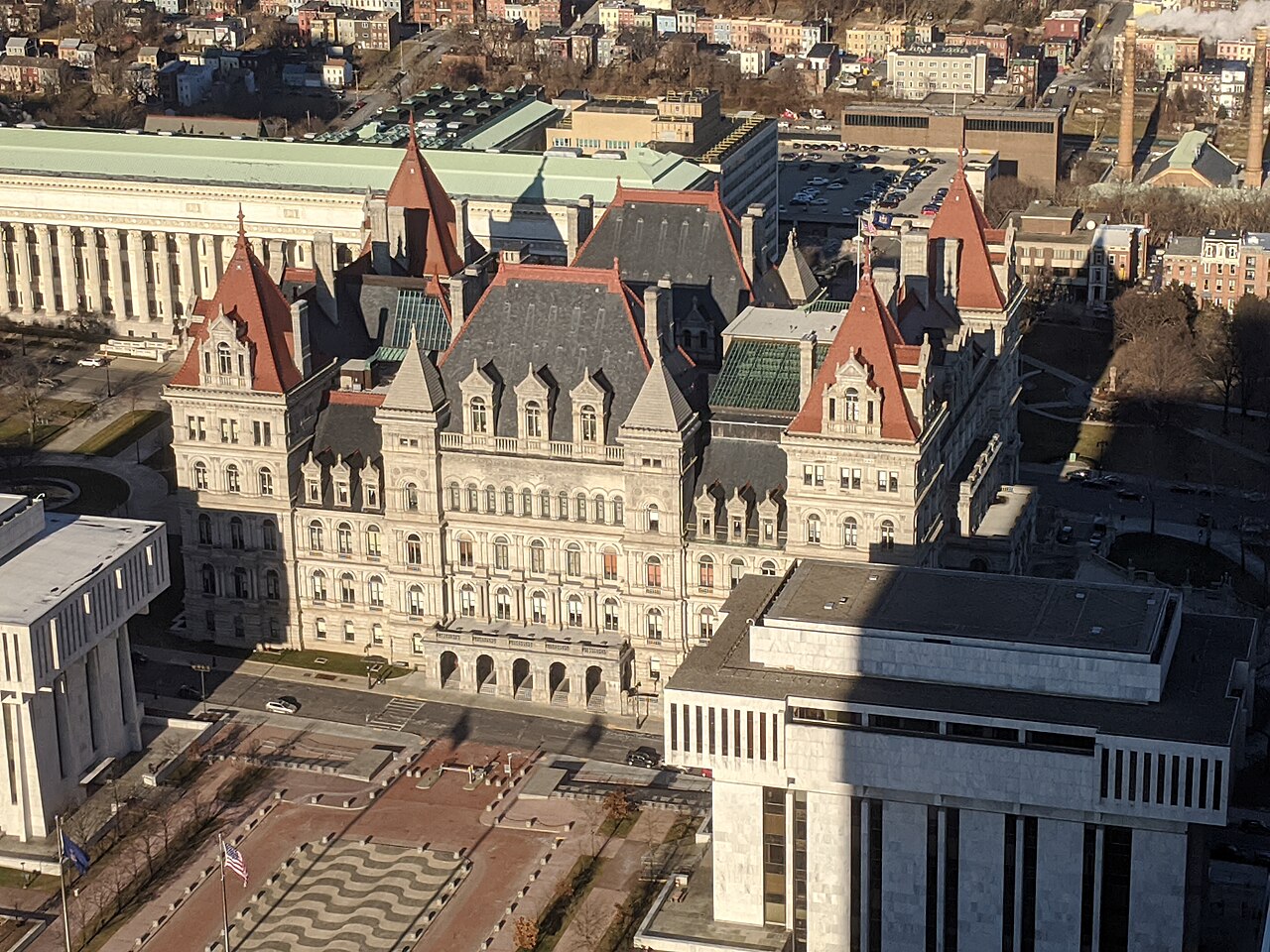 A large grey stone building with red pointed roofs and corner towers, seen from high above among city blocks.