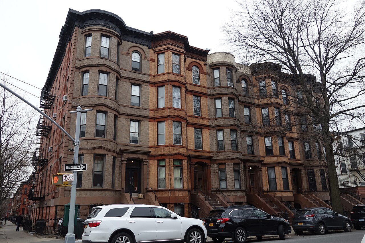 A row of four-storey brown stone houses with rounded bay fronts on a street corner, bare trees beside them.