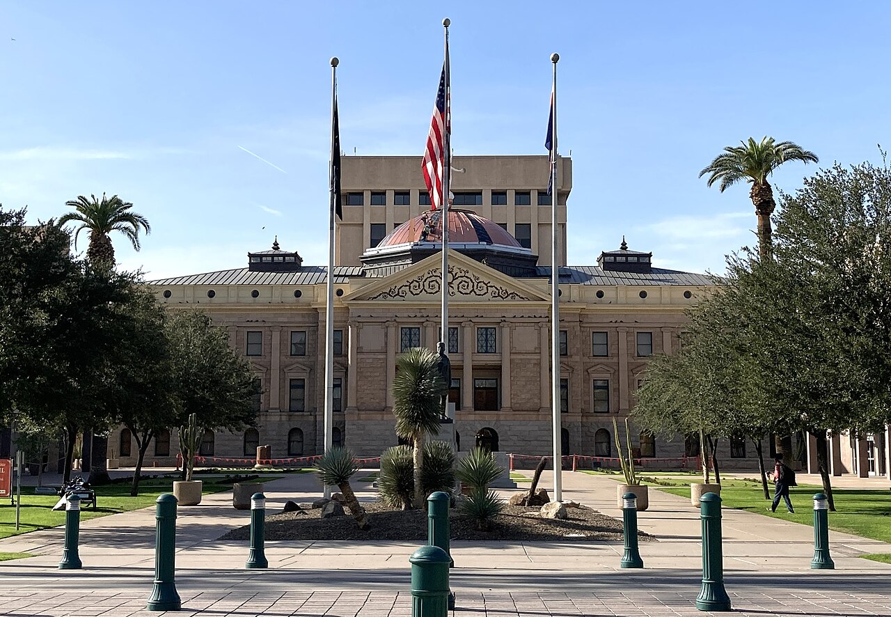 A low stone building with a small copper dome in front of a taller office block, with flagpoles and palm trees.