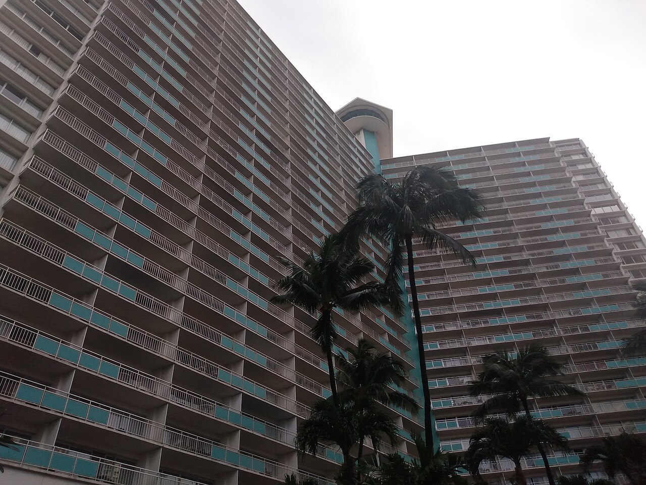 A wide tower with long rows of balconies seen from the ground, with palm trees in front.