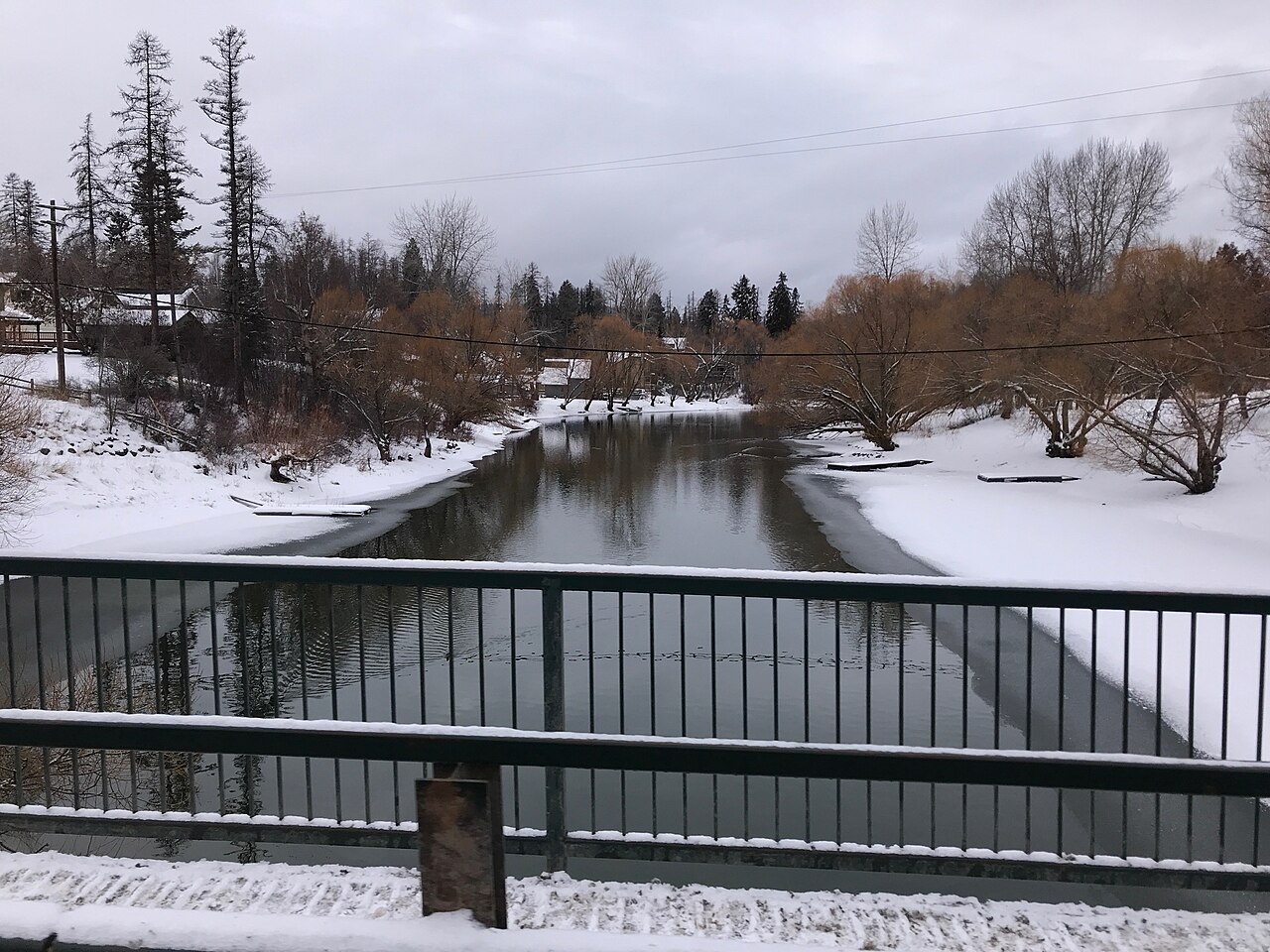 A dark river winding between snow-covered banks and bare trees, viewed over a bridge railing.