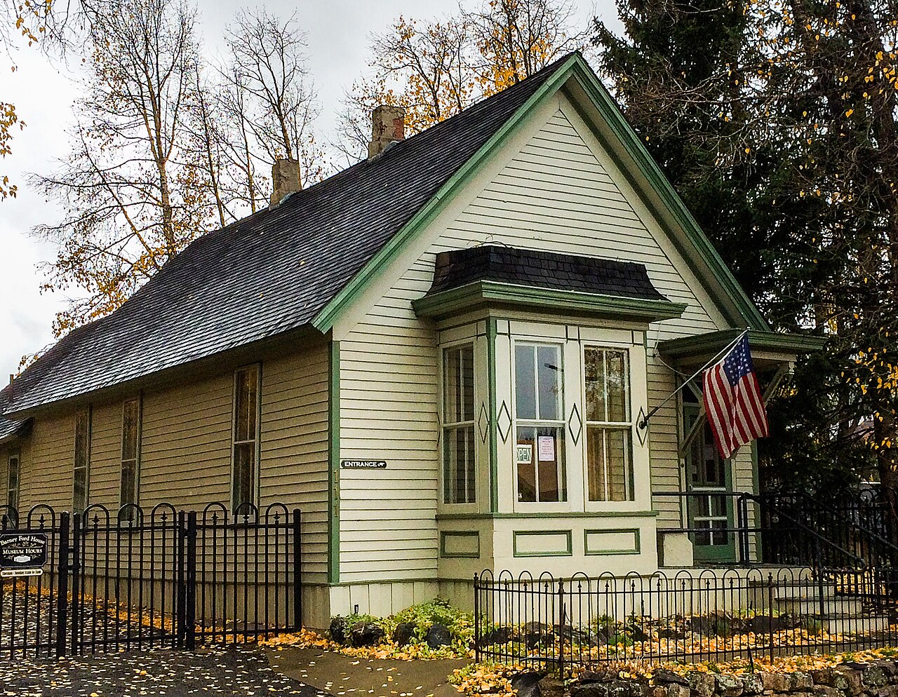 A small pale green wooden house with a bay window and a flag, behind an iron fence among autumn trees.