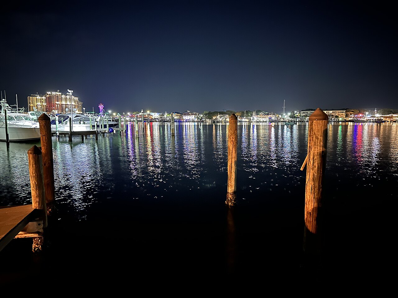 Coloured lights from a far shore reflected in dark water, with wooden mooring posts in the foreground.