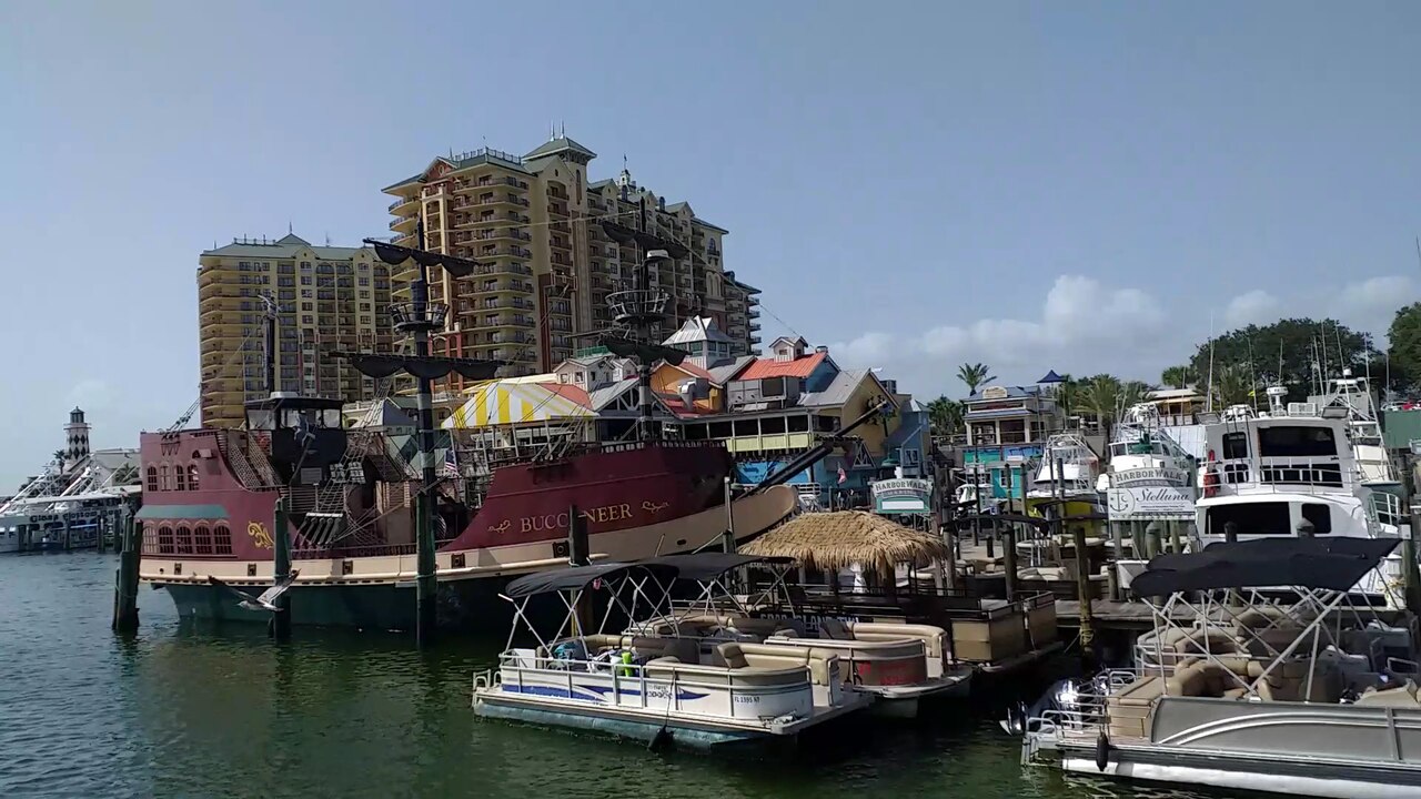 Pontoon boats and a red sailing ship moored below tall apartment towers at a busy marina.