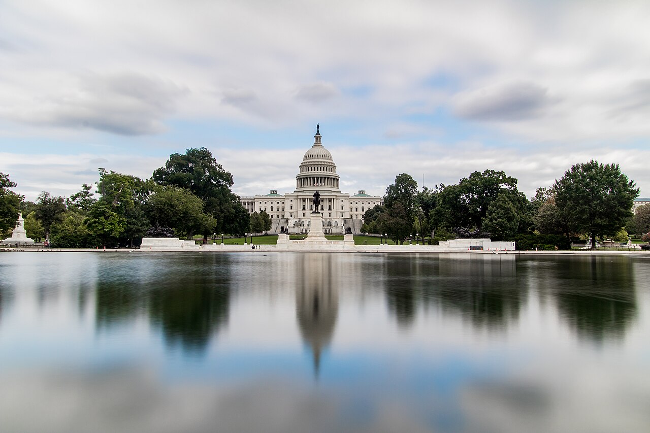 A white domed building framed by trees and mirrored in smooth water under a cloudy sky.