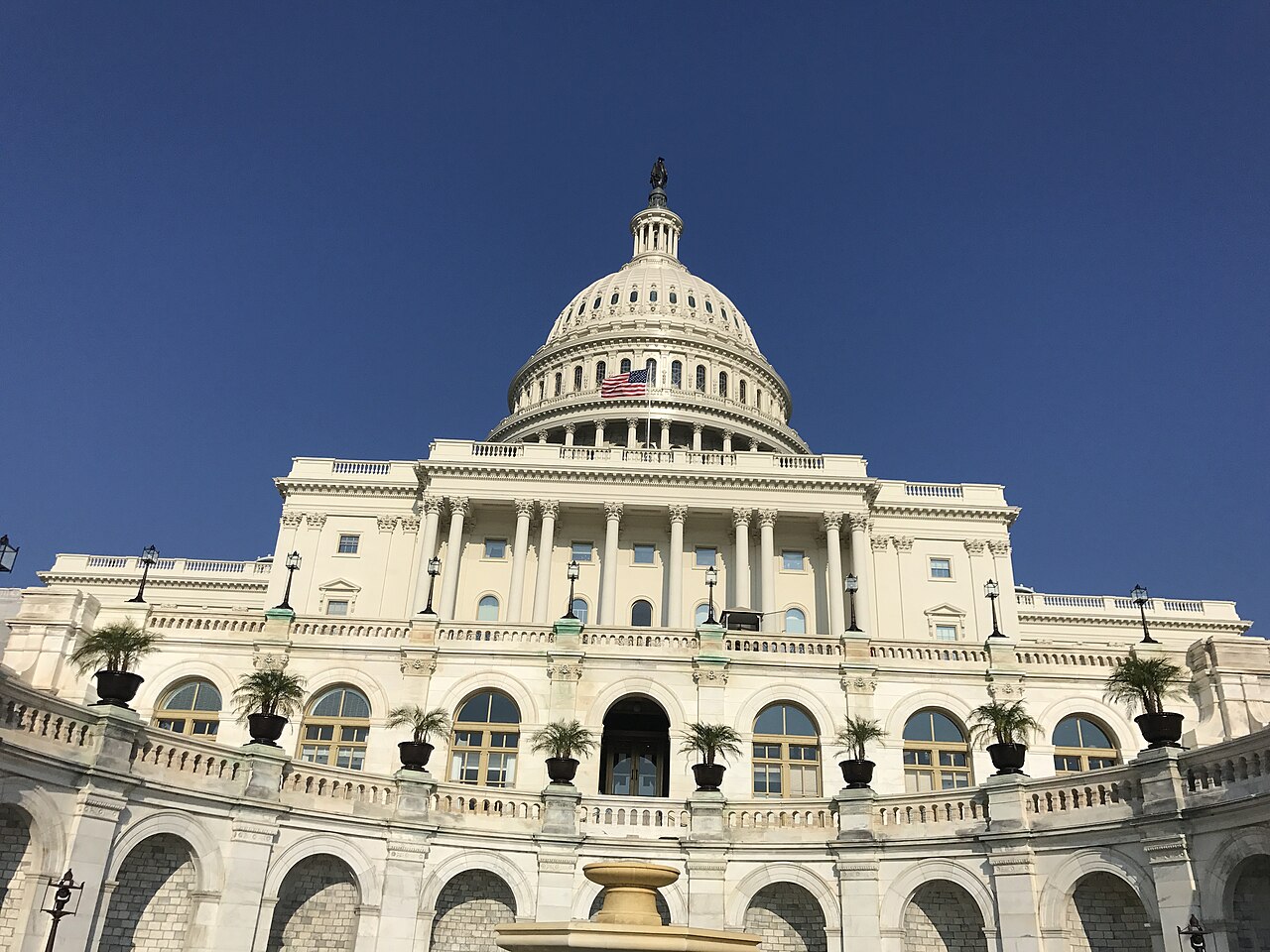 The white dome and columned front of a large building in bright sun against a dark blue sky.