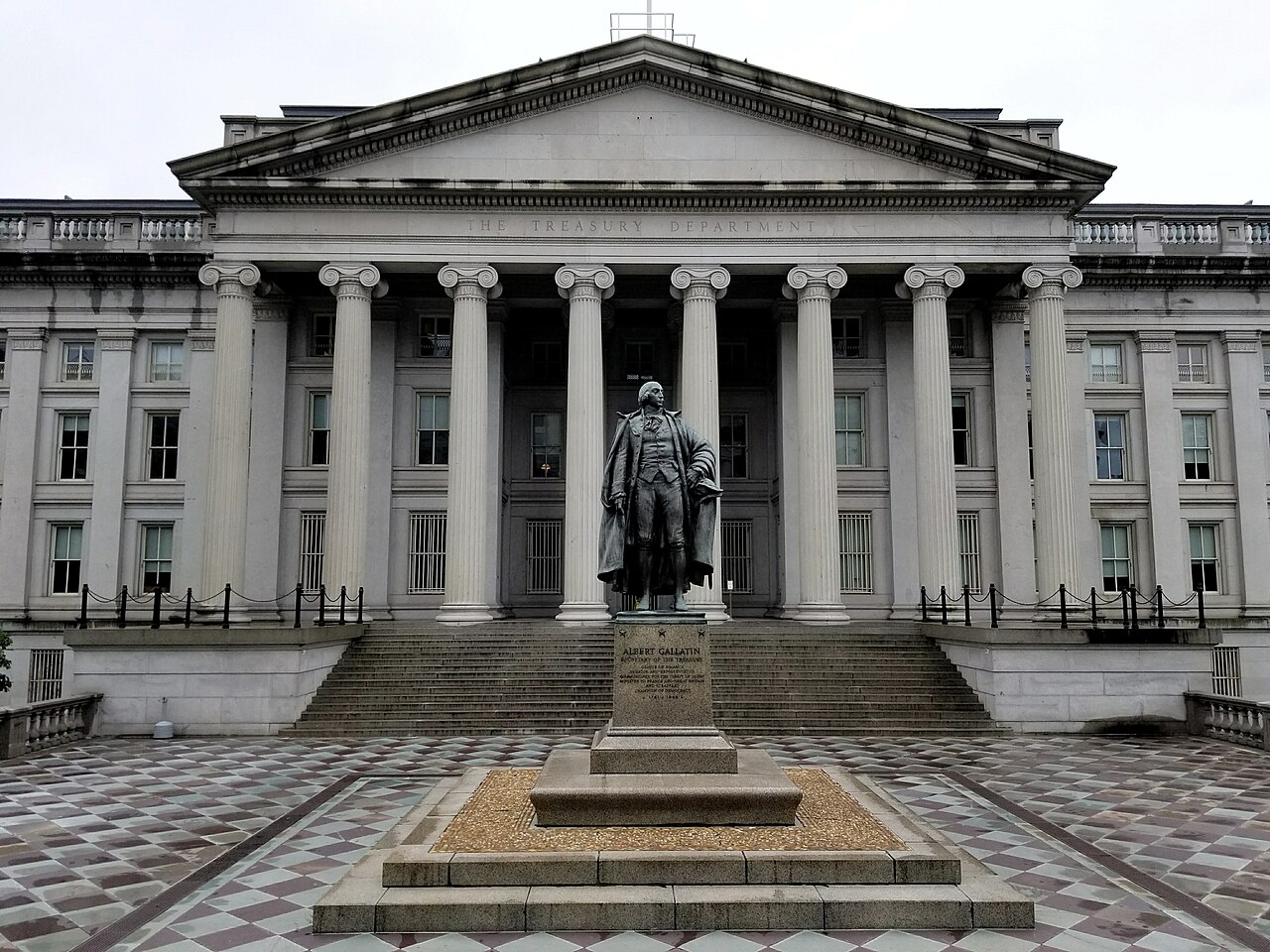 A grey stone building with tall columns and a bronze statue on a plinth in front of its steps.