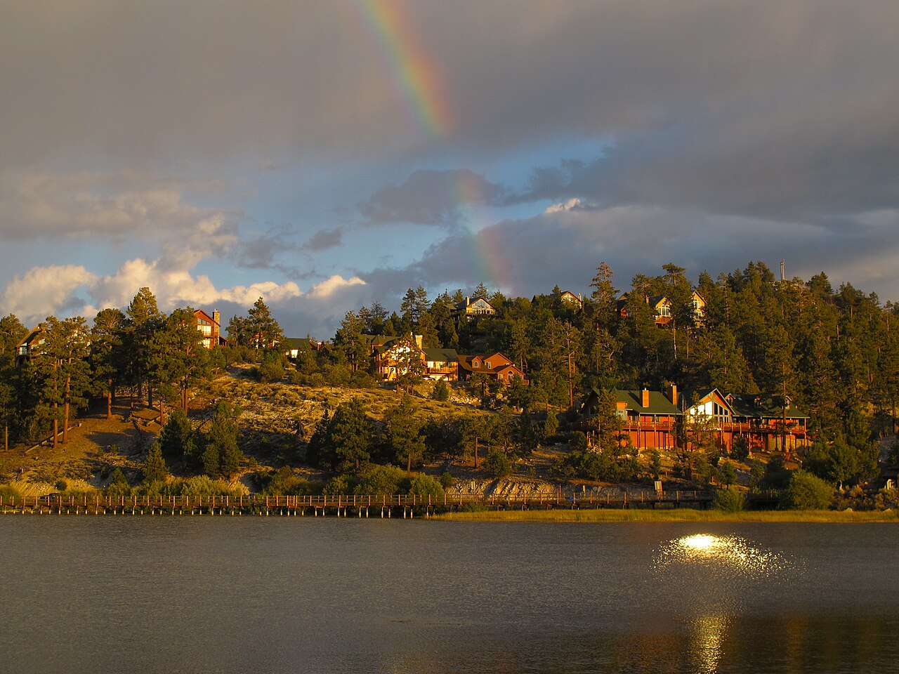 Houses on a wooded slope above a lake in warm evening light, with a faint rainbow in a grey sky.