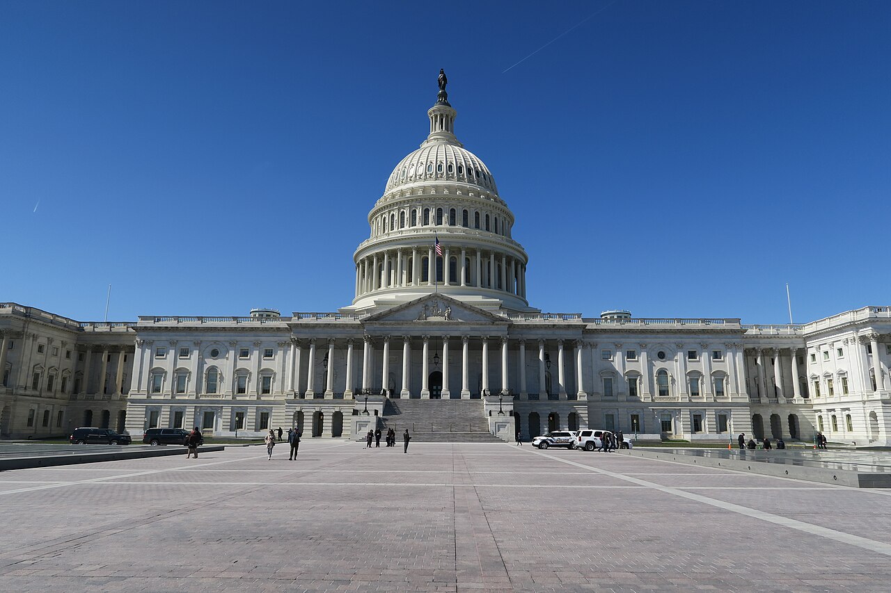 A white domed building seen straight on across a wide paved plaza under a clear blue sky.