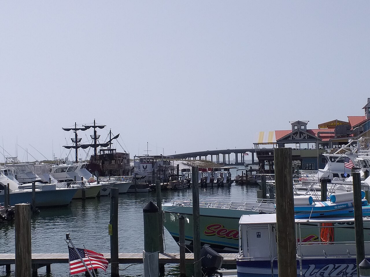 Fishing and pleasure boats at wooden docks, with a tall-masted ship and a road bridge behind.