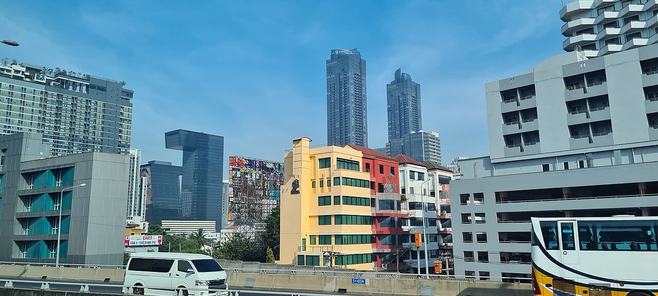 Two tall dark glass towers and a G-shaped office block rise behind low, brightly painted buildings under a blue sky.