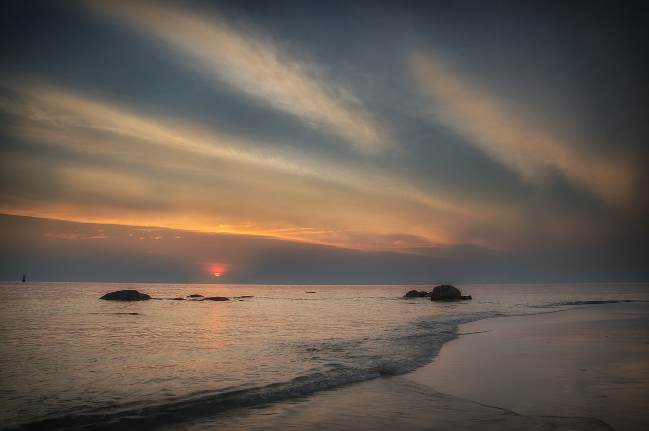 A low orange sun on the horizon under streaked cloud, with small rocks offshore and waves running up a dark beach.