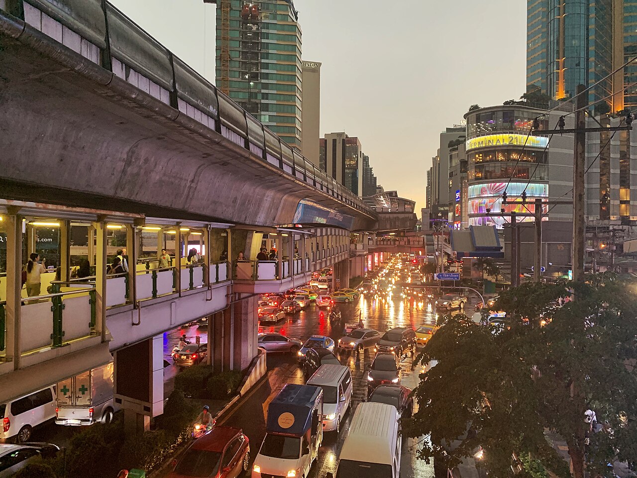 An elevated railway station runs above a wide road full of evening traffic, with towers and lit signs on both sides.