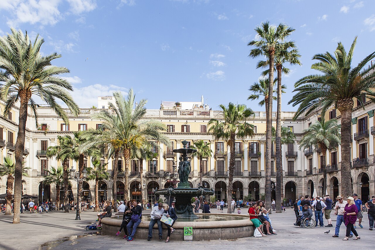 A square ringed by arcaded cream buildings, with tall palm trees and a fountain at its centre and people sitting around it.