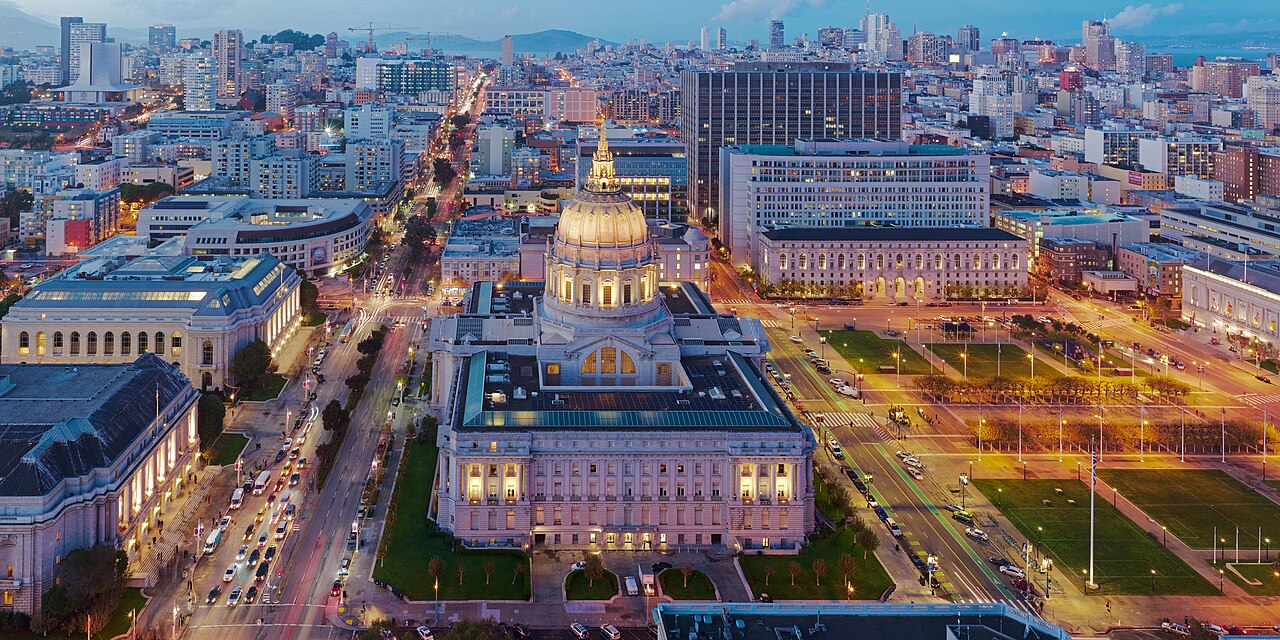 A domed civic building lit gold at dusk, seen from above with city streets and traffic lights spreading out around it.