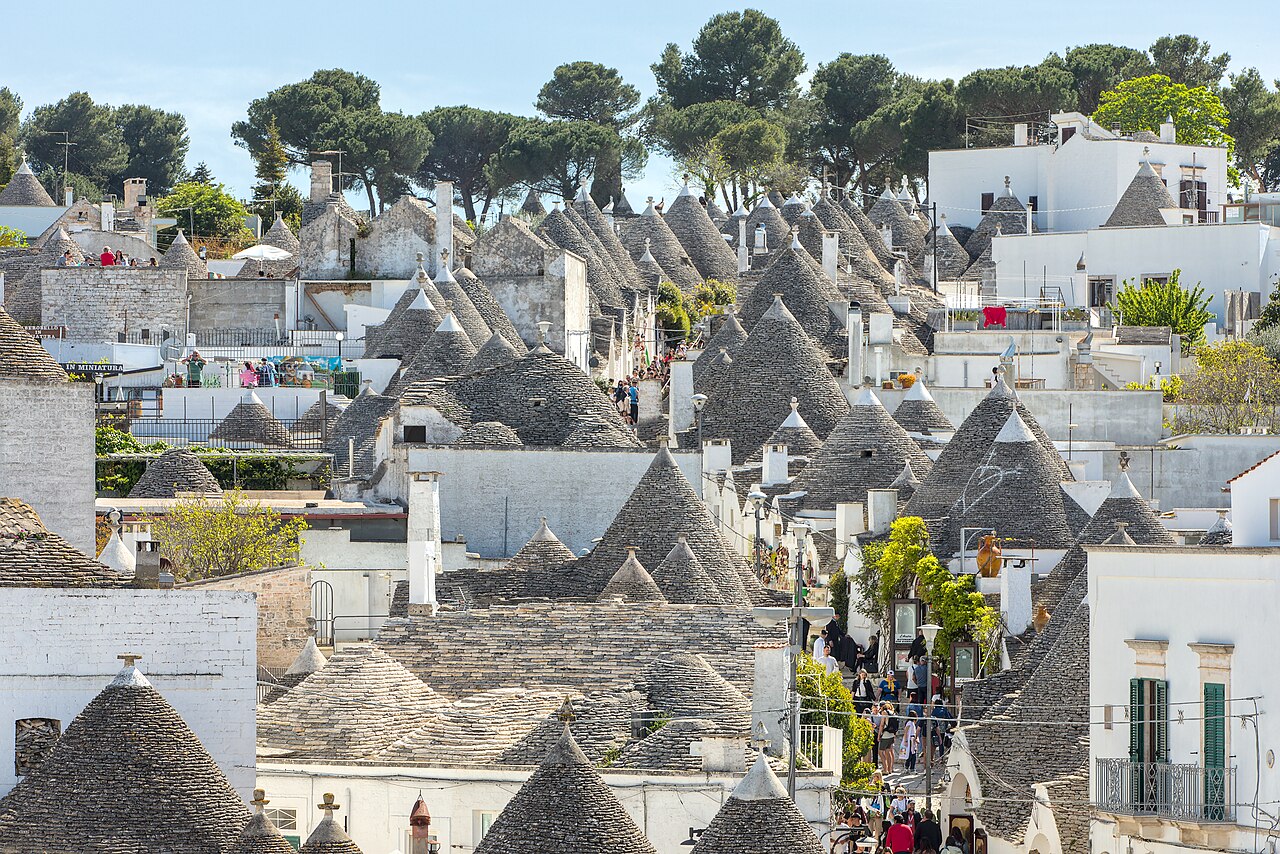 A hillside packed with small whitewashed houses, each topped with a grey conical stone roof, under pine trees.