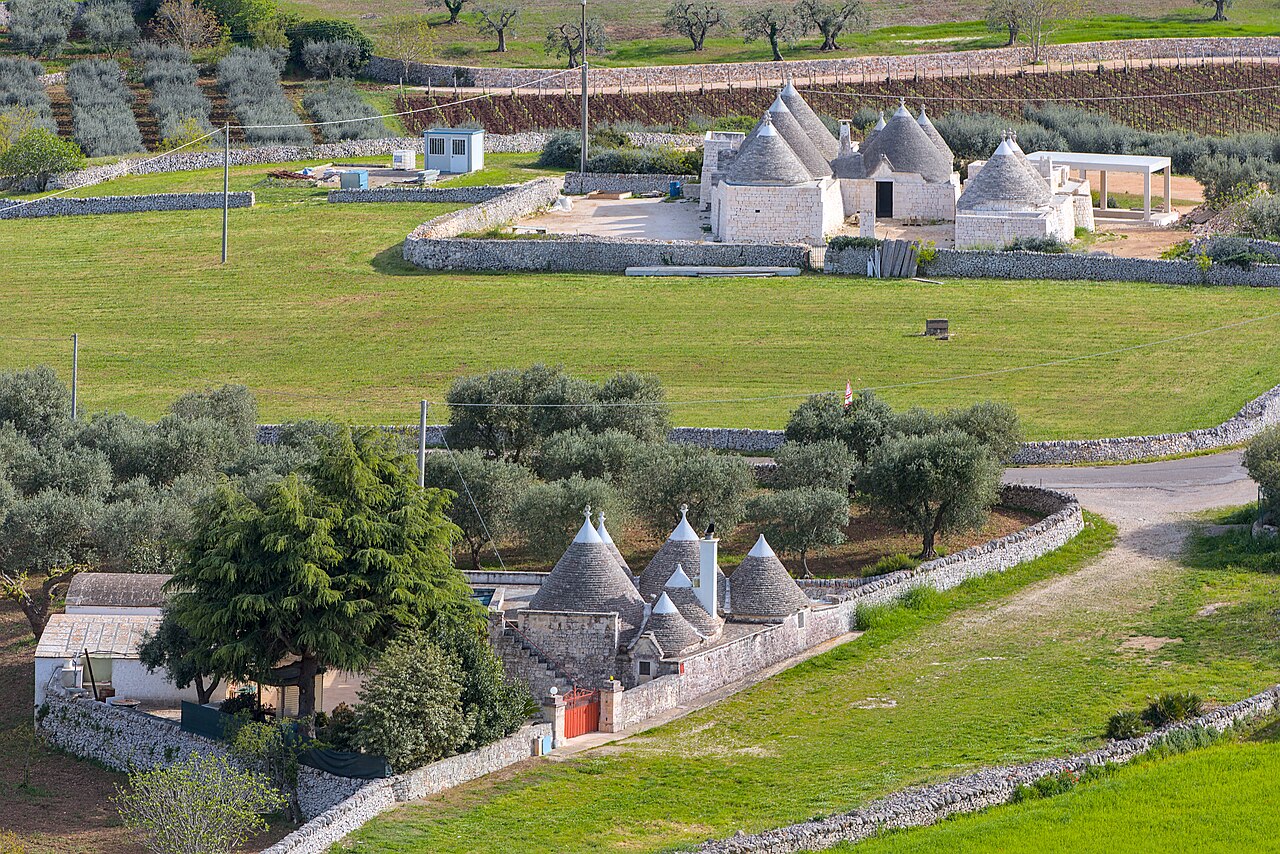 Two clusters of cone-roofed stone houses sit among green fields, olive trees and dry-stone walls, seen from above.