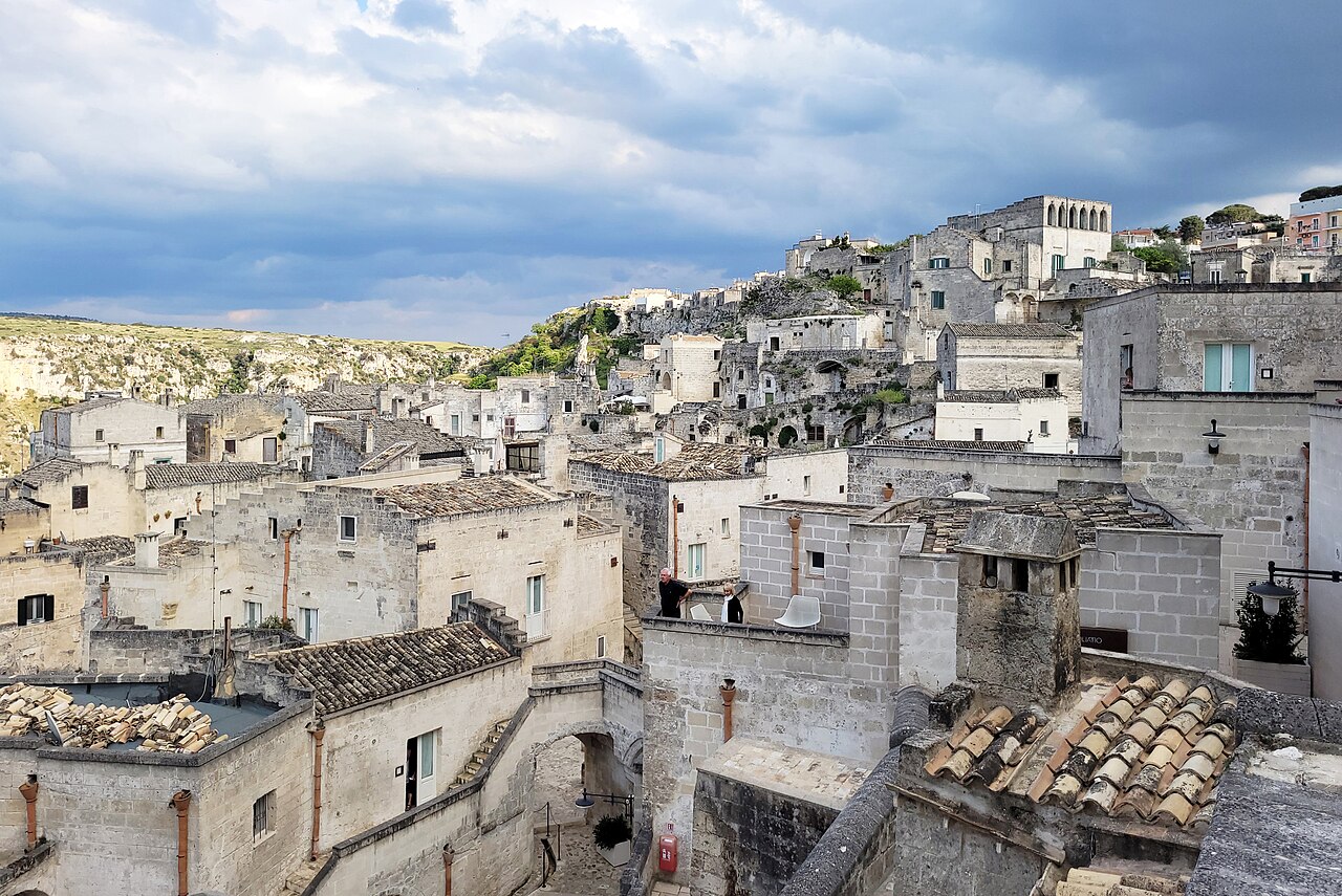 Pale stone houses stacked up a slope above a ravine, with tiled roofs in the foreground and heavy cloud overhead.