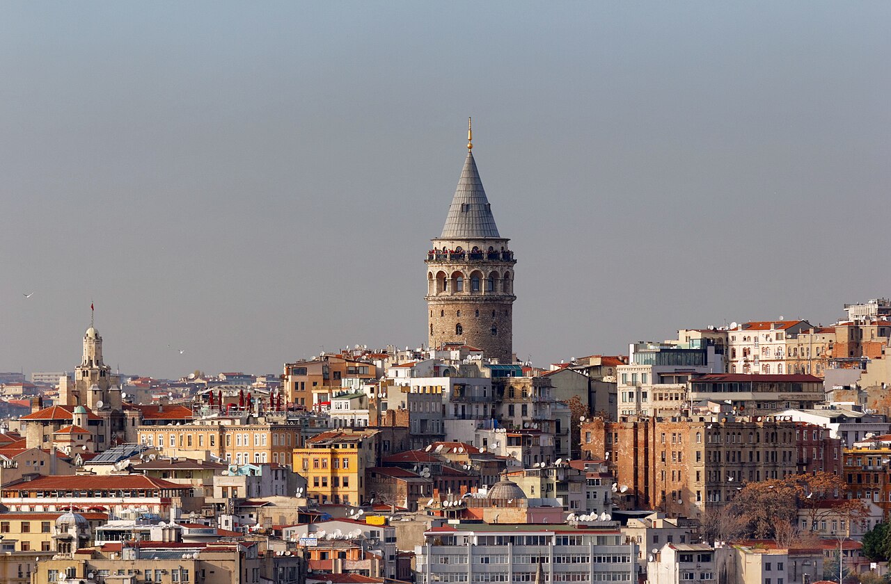 A round stone tower with a conical roof stands above a dense hillside of older apartment buildings in soft light.