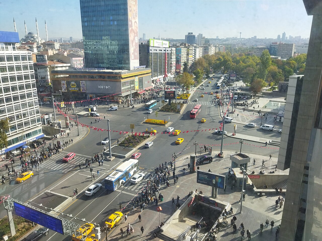 A busy city junction seen from a high window, with buses, yellow taxis, crowds on the pavements and office blocks around it.