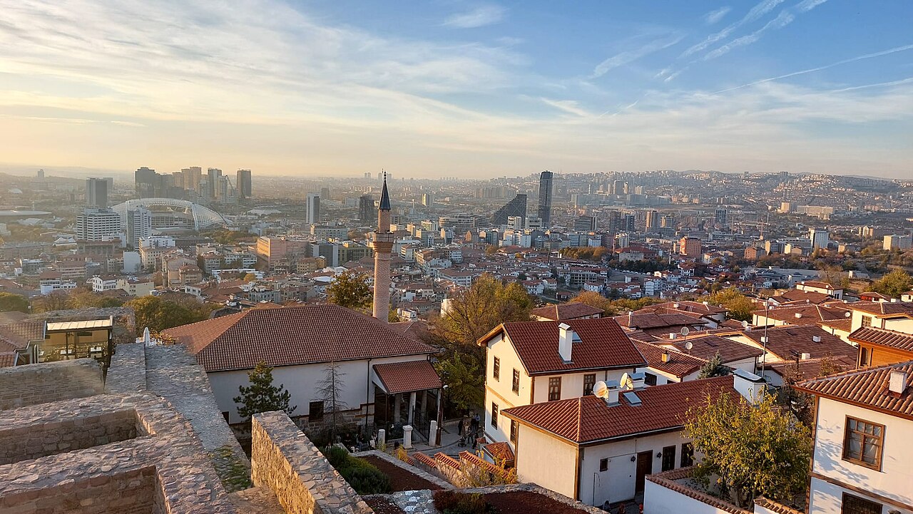 Red-tiled roofs and a slim minaret in the foreground, with a wide modern city stretching to hills under a pale evening sky.