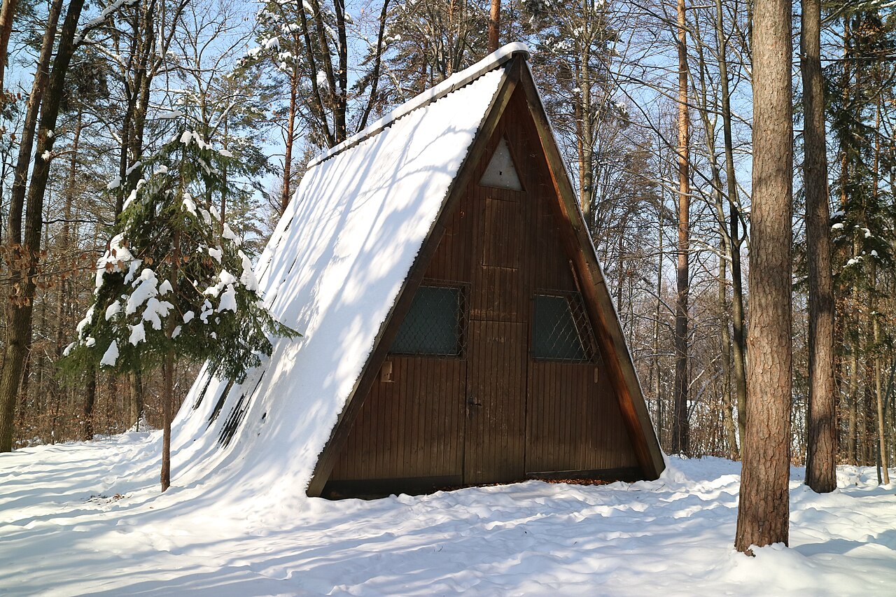 A small wooden house whose steep roof reaches the ground on both sides, half covered in snow among bare trees.