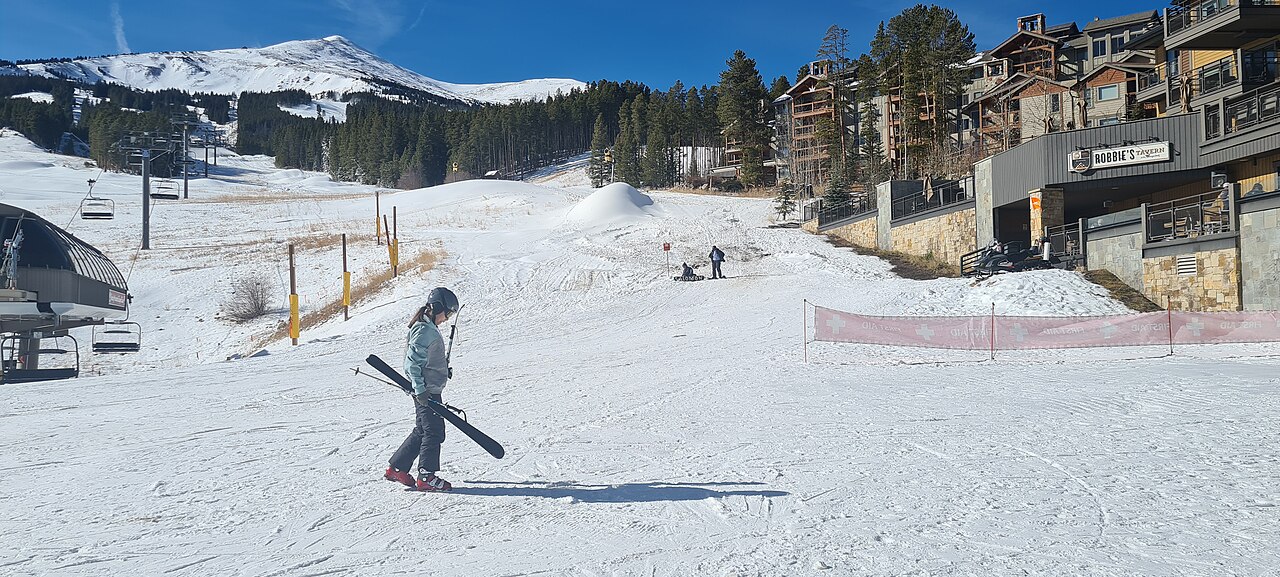 A snow-covered ski slope with lodge buildings and pine trees along its edge and a skier carrying skis across the foreground.