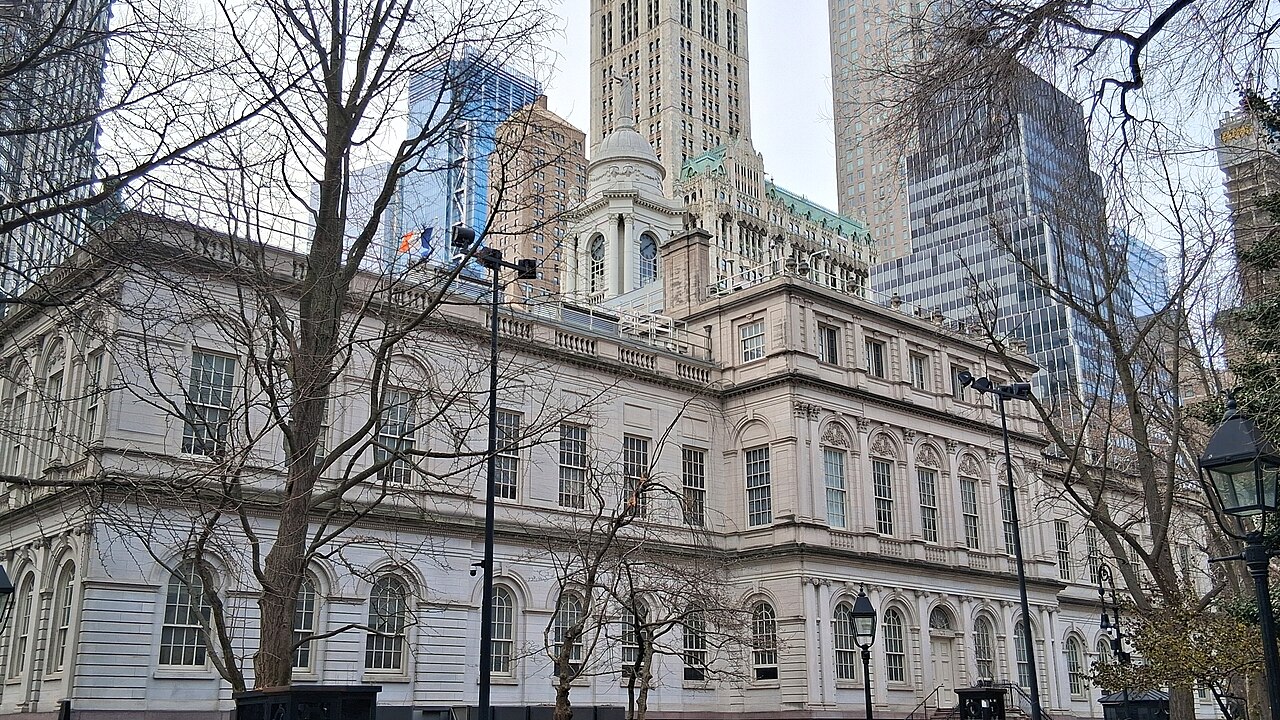 A pale stone civic building with arched windows and a cupola behind bare winter trees, with office towers rising behind it.