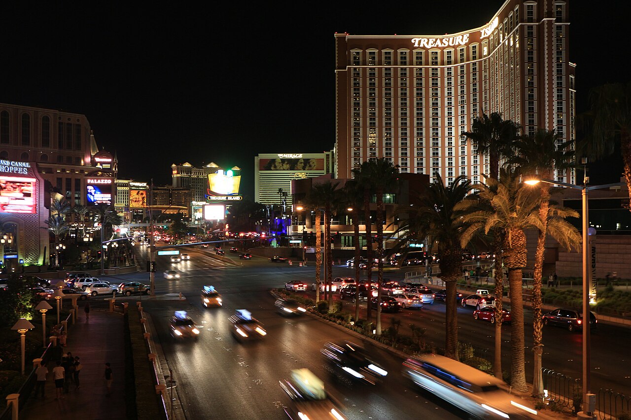 Light trails from traffic on a wide boulevard lined with palm trees, with lit hotel towers behind against a dark sky.