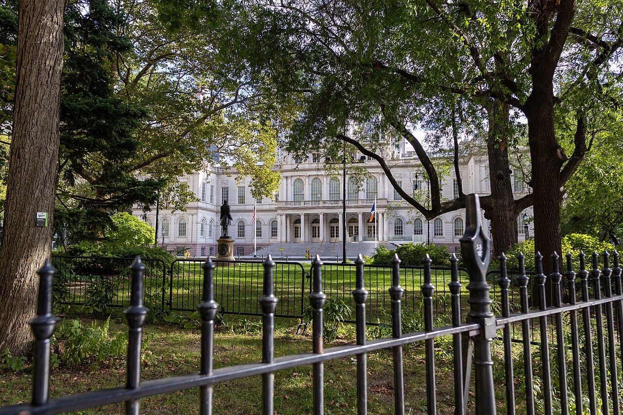 A pale stone building glimpsed across a lawn through leafy trees, behind a black iron fence in the foreground.