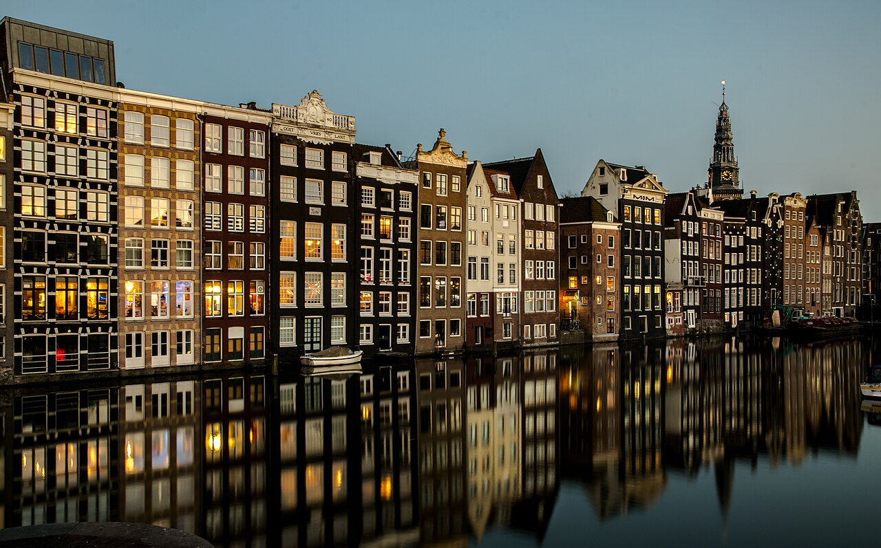 A row of narrow gabled houses with lit windows standing directly on still water that mirrors them, a church spire behind.