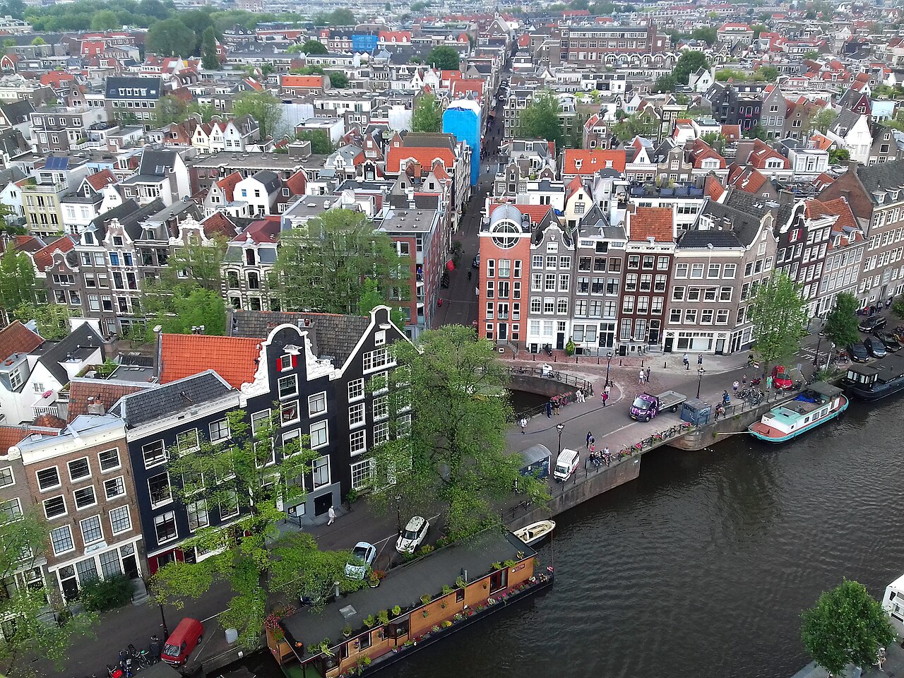 Rows of gabled brick houses with red and grey roofs packed along a tree-lined canal with moored houseboats, seen from above.