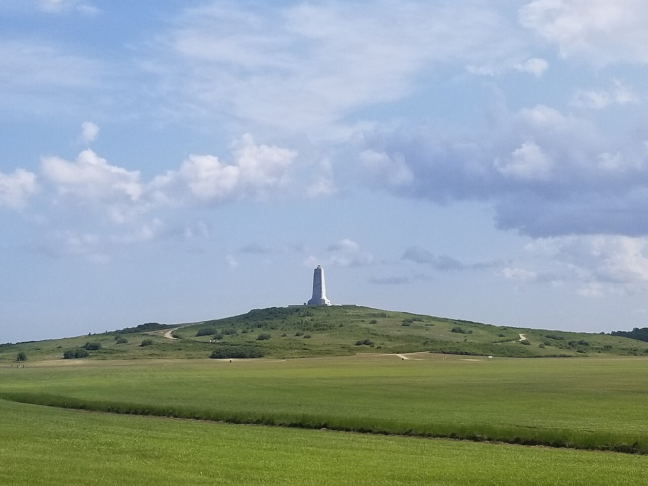 A stone pylon standing on top of a rounded green hill above a wide mown field, under a sky of scattered clouds.